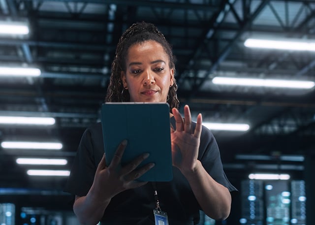 A woman in a dark industrial-style room using a tablet, possibly managing cybersecurity or IT operations.