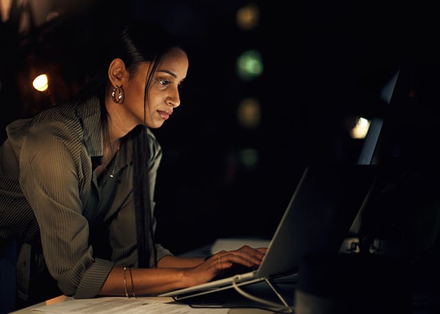 A woman working on a laptop in a dimly lit environment, focused on cybersecurity or data analysis.