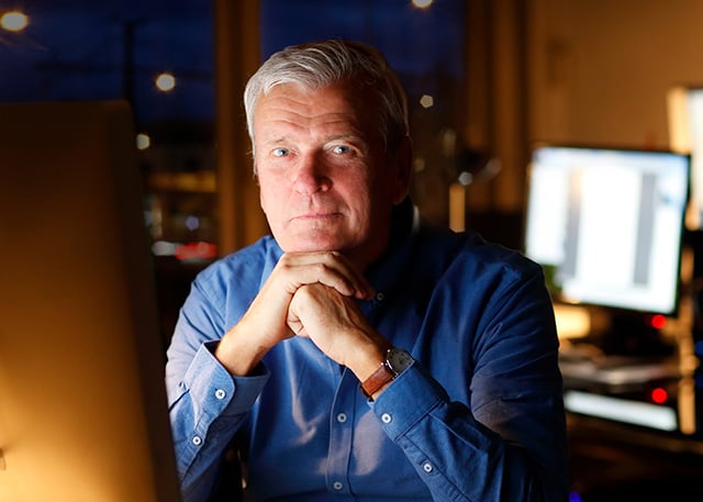 A confident older man in a blue shirt sitting at a desk in a dimly lit office, with computer screens in the background, symbolizing experience in cybersecurity or IT leadership.