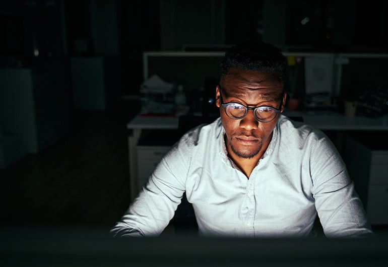 A focused man wearing glasses and a light-colored shirt working late at night in a dimly lit office, illuminated by his computer screen, symbolizing cybersecurity, coding, or IT operations