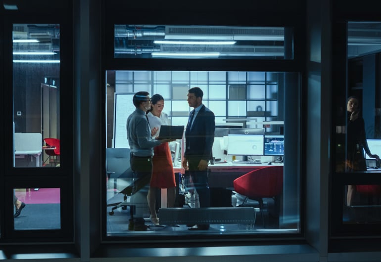 View through an office window at night showing three professionals collaborating around a laptop, with desks, monitors, and office equipment in the background, and another person working alone in a separate room.